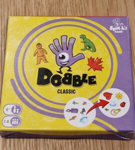 Image of the "Dobble Classic" card game box featuring colorful graphics, including a hand symbol, various icons like a dinosaur and a maple leaf, and game details indicating it's suitable for 2-8 players aged 4 and up. The box design includes a vibrant yellow background with purple accents.