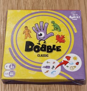 Image of the "Dobble Classic" card game box featuring colorful graphics, including a hand symbol, various icons like a dinosaur and a maple leaf, and game details indicating it's suitable for 2-8 players aged 4 and up. The box design includes a vibrant yellow background with purple accents.