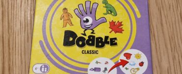 Image of the "Dobble Classic" card game box featuring colorful graphics, including a hand symbol, various icons like a dinosaur and a maple leaf, and game details indicating it's suitable for 2-8 players aged 4 and up. The box design includes a vibrant yellow background with purple accents.