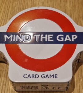 Mind the Gap card game in a round tin box with a red and white London Underground logo, resting on a wooden surface.