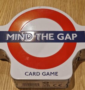 Mind the Gap card game in a round tin box with a red and white London Underground logo, resting on a wooden surface.