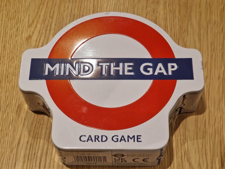 Mind the Gap card game in a round tin box with a red and white London Underground logo, resting on a wooden surface.