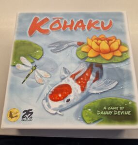 Colorful board game box for "Kōhaku" designed by Danny Devine, featuring a koi fish, a water lily, and dragonflies, highlighting its serene aquatic theme.