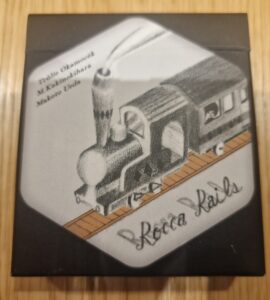 Illustration of a vintage steam locomotive on the packaging of the board game "Rocca Rails," featuring the names of the designers: Tsubasa Okamoto, M. Kakinohihara, and Makoto Ueda.