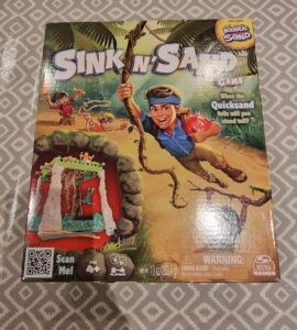 Box cover of the "Sink n' Sand" game featuring vibrant illustrations of children playing in a jungle setting, with the main character hanging from a vine. The game includes kinetic sand and is designed for 2-4 players aged 4 and up. The packaging highlights the fun challenge of balancing as quicksand falls.