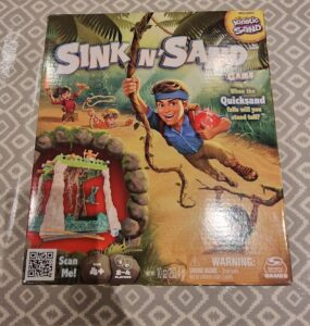 Box cover of the "Sink n' Sand" game featuring vibrant illustrations of children playing in a jungle setting, with the main character hanging from a vine. The game includes kinetic sand and is designed for 2-4 players aged 4 and up. The packaging highlights the fun challenge of balancing as quicksand falls.