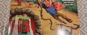 Box cover of the "Sink n' Sand" game featuring vibrant illustrations of children playing in a jungle setting, with the main character hanging from a vine. The game includes kinetic sand and is designed for 2-4 players aged 4 and up. The packaging highlights the fun challenge of balancing as quicksand falls.