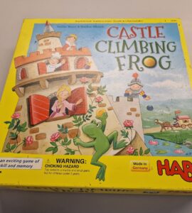 Colorful box of the board game "Castle Climbing Frog" by HABA, featuring playful illustrations of a frog, castle, and characters. The bright yellow packaging highlights the game’s focus on skill and memory, with a warning label for small parts. Made in Germany.
