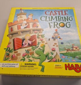 Colorful box of the board game "Castle Climbing Frog" by HABA, featuring playful illustrations of a frog, castle, and characters. The bright yellow packaging highlights the game’s focus on skill and memory, with a warning label for small parts. Made in Germany.