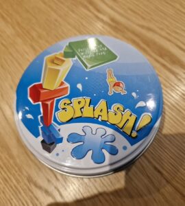 Colorful game tin for "Splash!" featuring playful graphics and a blue splash design, placed on a wooden table surface.