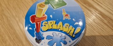 Colorful game tin for "Splash!" featuring playful graphics and a blue splash design, placed on a wooden table surface.
