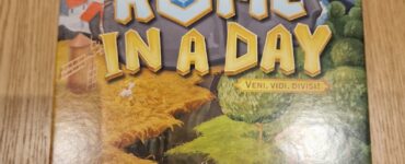 Board game "Rome in a Day" featuring vibrant artwork of ancient Rome, with the tagline "Veni, Vidi, Divisi," designed by Evgeny Petrov, published by Red Cat Games. The cover showcases a picturesque landscape with buildings, greenery, and a horse, set against a backdrop of the iconic Colosseum.