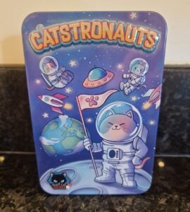 Colorful illustration of a tin featuring "Catstronauts," depicting cute cats in astronaut suits exploring space, with planets and a flag on the moon. Ideal for cat lovers and space enthusiasts.