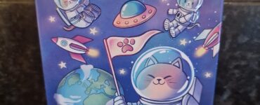 Colorful illustration of a tin featuring "Catstronauts," depicting cute cats in astronaut suits exploring space, with planets and a flag on the moon. Ideal for cat lovers and space enthusiasts.