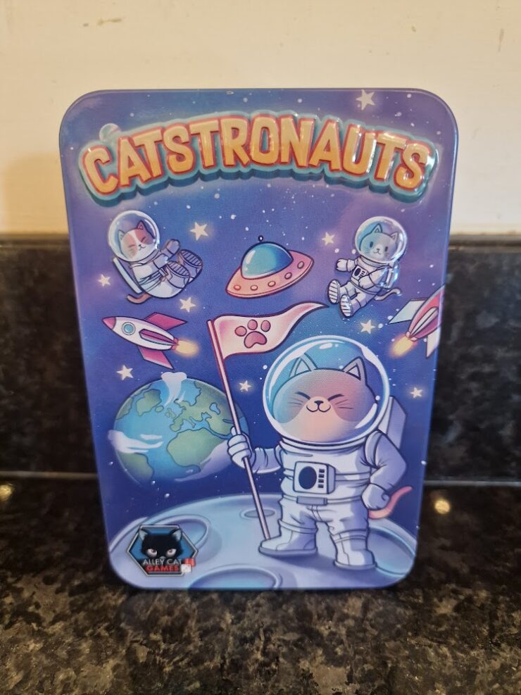 Colorful illustration of a tin featuring "Catstronauts," depicting cute cats in astronaut suits exploring space, with planets and a flag on the moon. Ideal for cat lovers and space enthusiasts.