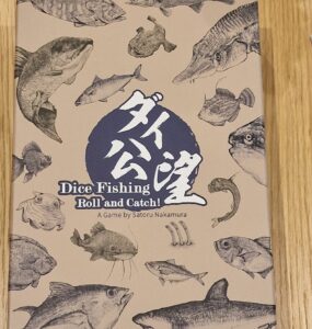 Dice Fishing game box featuring various fish illustrations, titled "Dice Fishing: Roll and Catch!" designed by Satoru Nakamura, set against a light brown background.