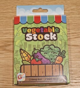 Colorful packaging of vegetable stock featuring cartoon vegetables like corn, tomatoes, and broccoli, on a wooden table. The product is branded as "Vegetable Stock" and is associated with a game called "Zong-Ger."