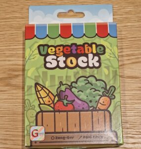 Colorful packaging of vegetable stock featuring cartoon vegetables like corn, tomatoes, and broccoli, on a wooden table. The product is branded as "Vegetable Stock" and is associated with a game called "Zong-Ger."