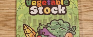 Colorful packaging of vegetable stock featuring cartoon vegetables like corn, tomatoes, and broccoli, on a wooden table. The product is branded as "Vegetable Stock" and is associated with a game called "Zong-Ger."
