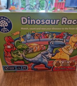 Colorful box of the "Dinosaur Race" game by Orchard Toys, featuring cartoon dinosaurs and a racetrack with medals. The game encourages matching, counting, and racing, suitable for ages 3-6 and designed for 2-4 players.