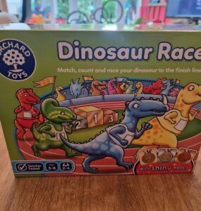 Colorful box of the "Dinosaur Race" game by Orchard Toys, featuring cartoon dinosaurs and a racetrack with medals. The game encourages matching, counting, and racing, suitable for ages 3-6 and designed for 2-4 players.