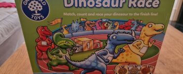 Colorful box of the "Dinosaur Race" game by Orchard Toys, featuring cartoon dinosaurs and a racetrack with medals. The game encourages matching, counting, and racing, suitable for ages 3-6 and designed for 2-4 players.