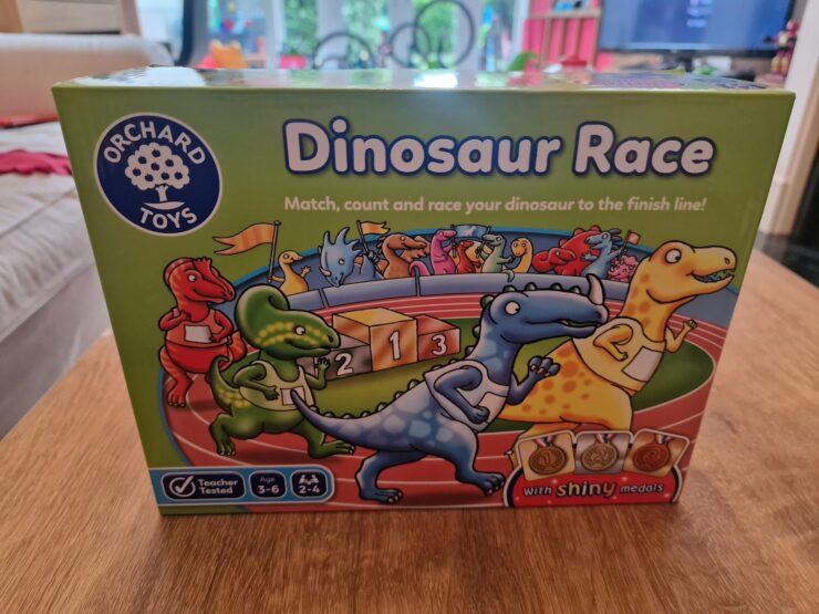 Colorful box of the "Dinosaur Race" game by Orchard Toys, featuring cartoon dinosaurs and a racetrack with medals. The game encourages matching, counting, and racing, suitable for ages 3-6 and designed for 2-4 players.