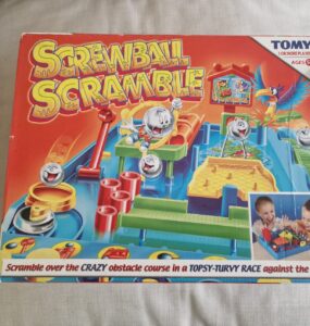 Colorful box of the game "Screwball Scramble" by TOMY featuring a crazy obstacle course for one or more players. Suitable for ages 5 and up, the game challenges players to navigate balls through various obstacles in a timed race. The box shows children engaged in gameplay, highlighting the fun and competitive nature of the game.