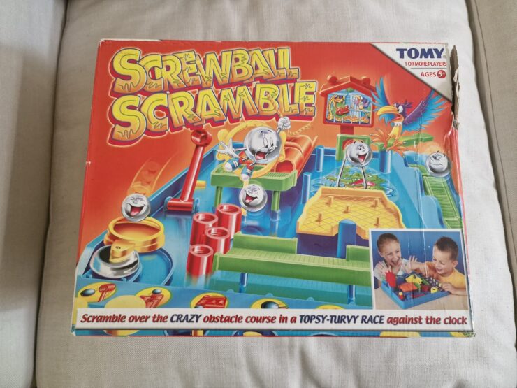 Colorful box of the game "Screwball Scramble" by TOMY featuring a crazy obstacle course for one or more players. Suitable for ages 5 and up, the game challenges players to navigate balls through various obstacles in a timed race. The box shows children engaged in gameplay, highlighting the fun and competitive nature of the game.