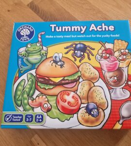 Colorful game box for "Tummy Ache" by Orchard Toys, featuring a cartoon hamburger, vegetables, and desserts with playful insects. Suitable for ages 3-7, this fun family game encourages children to create meals while avoiding yucky foods.