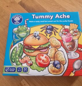 Colorful game box for "Tummy Ache" by Orchard Toys, featuring a cartoon hamburger, vegetables, and desserts with playful insects. Suitable for ages 3-7, this fun family game encourages children to create meals while avoiding yucky foods.