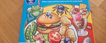 Colorful game box for "Tummy Ache" by Orchard Toys, featuring a cartoon hamburger, vegetables, and desserts with playful insects. Suitable for ages 3-7, this fun family game encourages children to create meals while avoiding yucky foods.