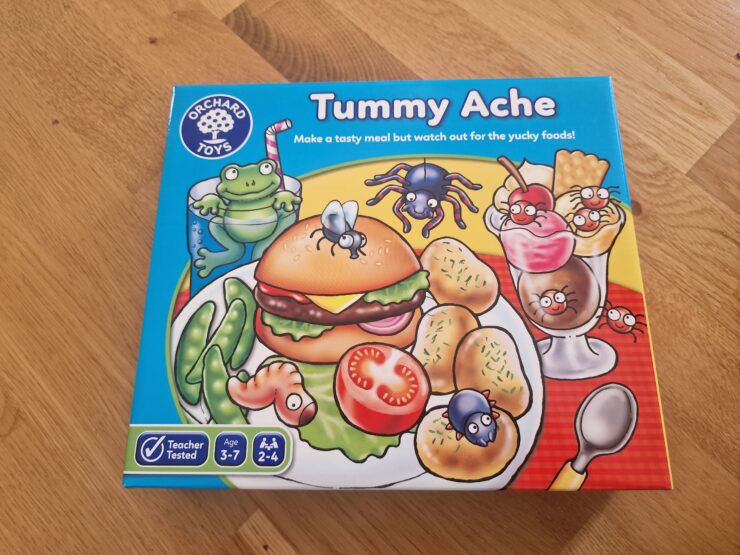 Colorful game box for "Tummy Ache" by Orchard Toys, featuring a cartoon hamburger, vegetables, and desserts with playful insects. Suitable for ages 3-7, this fun family game encourages children to create meals while avoiding yucky foods.