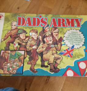 Box cover of the board game "Dad's Army," featuring colorful illustrations of characters in military attire, including a soldier with a rifle and others in various poses. The design includes the British flag and playful text, highlighting the game's theme of humor and strategy. Ideal for fans of classic British television and board games.