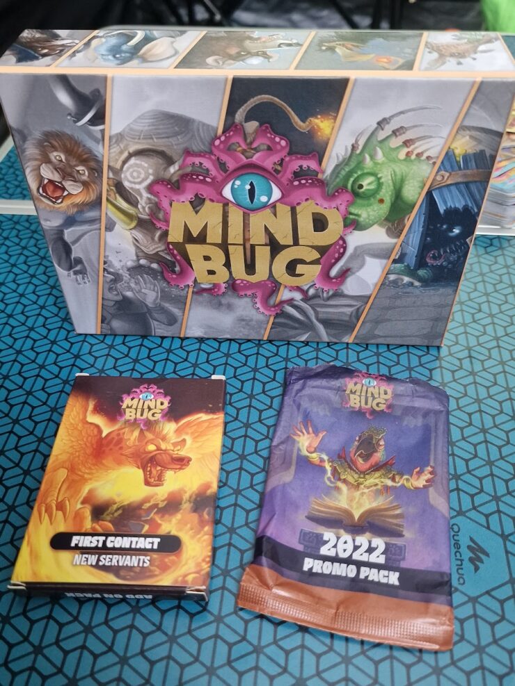 Box set of the card game "Mind Bug" featuring the main game box, a "First Contact" expansion pack, and a "2022 Promo Pack." The game combines strategy and fantasy elements, showcasing vibrant artwork of various characters and creatures.