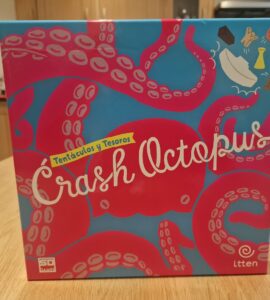Colorful board game box for "Crash Octopus," featuring playful octopus graphics and the subtitle "Tentáculos y Tesoros." The box is displayed on a wooden table with a kitchen background.