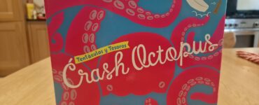 Colorful board game box for "Crash Octopus," featuring playful octopus graphics and the subtitle "Tentáculos y Tesoros." The box is displayed on a wooden table with a kitchen background.