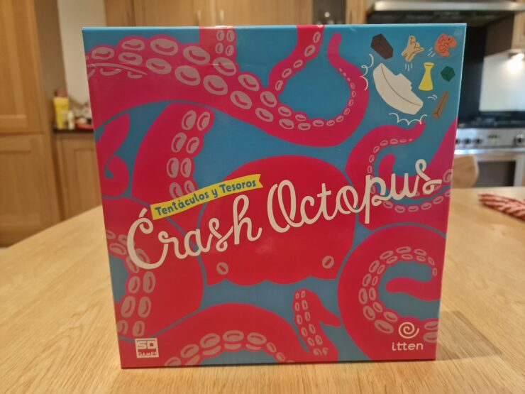 Colorful board game box for "Crash Octopus," featuring playful octopus graphics and the subtitle "Tentáculos y Tesoros." The box is displayed on a wooden table with a kitchen background.