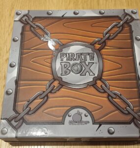 Pirate Box board game packaging featuring a wooden design with chains and a circular logo, designed for family-friendly gameplay and adventure-themed fun.