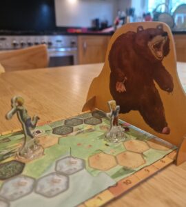 Board game scene featuring two player figures on a hexagonal game board, with a large bear cutout in the background. The setting is a cozy kitchen, highlighting a fun gaming experience.