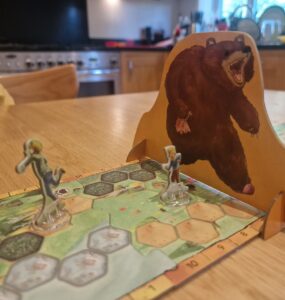 Board game scene featuring two player figures on a hexagonal game board, with a large bear cutout in the background. The setting is a cozy kitchen, highlighting a fun gaming experience.
