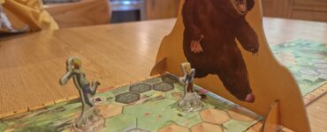 Board game scene featuring two player figures on a hexagonal game board, with a large bear cutout in the background. The setting is a cozy kitchen, highlighting a fun gaming experience.