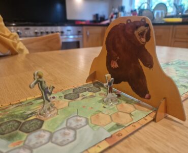 Board game scene featuring two player figures on a hexagonal game board, with a large bear cutout in the background. The setting is a cozy kitchen, highlighting a fun gaming experience.