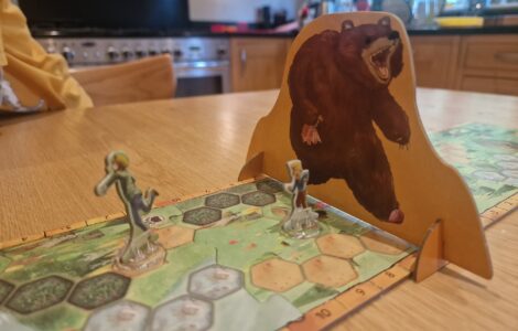 Board game scene featuring two player figures on a hexagonal game board, with a large bear cutout in the background. The setting is a cozy kitchen, highlighting a fun gaming experience.