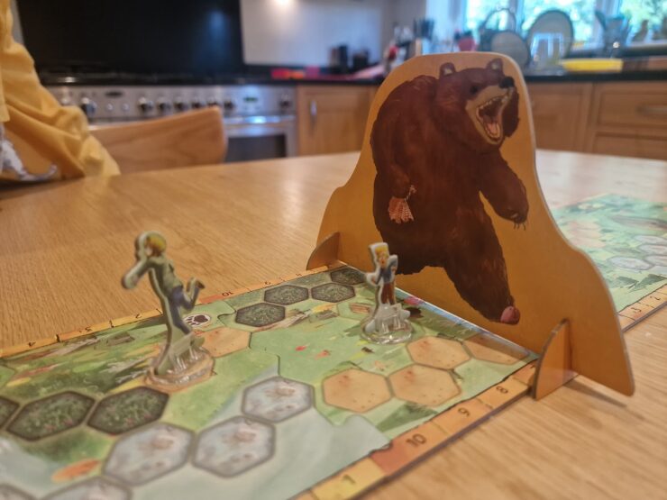 Board game scene featuring two player figures on a hexagonal game board, with a large bear cutout in the background. The setting is a cozy kitchen, highlighting a fun gaming experience.