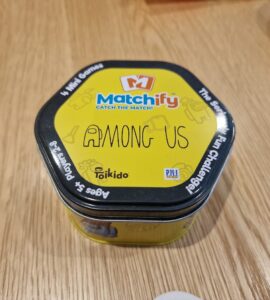 Alt text: A hexagonal tin container of the game "Matchify" featuring "Among Us," designed for 2-8 players aged 5 and up. The bright yellow tin showcases the game's title prominently, highlighting its fun and engaging mini-games.