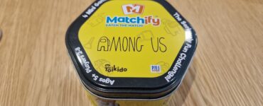 Alt text: A hexagonal tin container of the game "Matchify" featuring "Among Us," designed for 2-8 players aged 5 and up. The bright yellow tin showcases the game's title prominently, highlighting its fun and engaging mini-games.