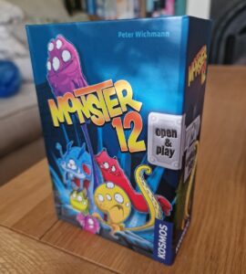Colorful board game box for "Monster 12" by Peter Wichmann, featuring playful monster illustrations and the text "open & play," designed for family-friendly fun. The box is placed on a wooden table with a cozy background.