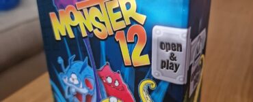Colorful board game box for "Monster 12" by Peter Wichmann, featuring playful monster illustrations and the text "open & play," designed for family-friendly fun. The box is placed on a wooden table with a cozy background.