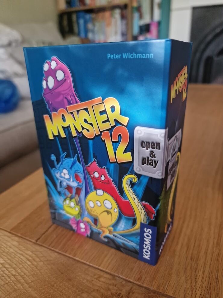 Colorful board game box for "Monster 12" by Peter Wichmann, featuring playful monster illustrations and the text "open & play," designed for family-friendly fun. The box is placed on a wooden table with a cozy background.